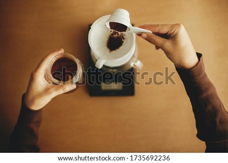 Hands pouring grounded coffee in filter pour over on glass kettle on scale,top view. Preparing for alternative coffee brewing. Person holding spoon with grind coffee on brown background Royalty-Free Stock Photo #1735692236