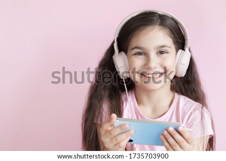 Cute little girl in headphones is using a smartphone, looking at camera and smiling on pastel background

