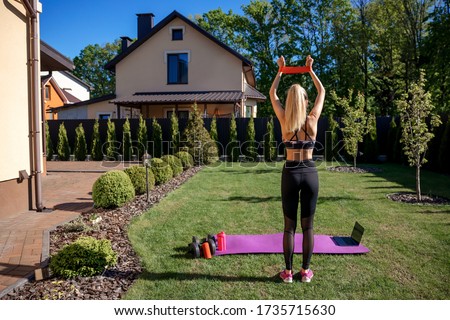 Pretty sportive fitness coach woman using  fitness gum in hand  at the garden with fresh green grass near house.  View from the back