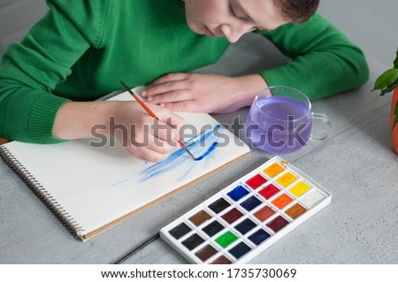 Close Up photo boy is painting with watercolors sitting at table, watercolor palette on grey background