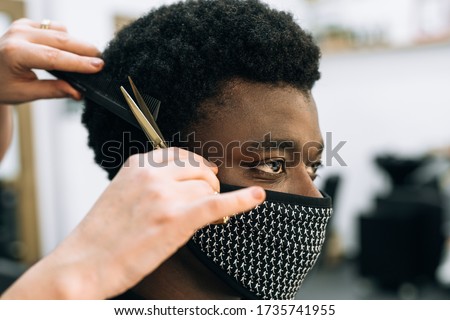 face of a black guy getting a haircut in a hair salon with a black mask on his face from the coronavirus. The hair is like afro. The scissors are golden. Royalty-Free Stock Photo #1735741955