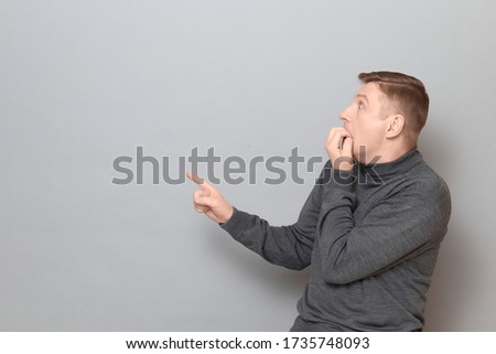 Studio shot of shocked frightened mature man wearing jumper, being full of fear, biting nails, pointing with finger at something awful, standing sideways over gray background, copy space on left