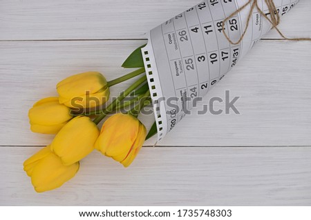 A natural bouquet of yellow tulips wrapped in a leaf from the calendar lies on a wooden white Board. Background, texture, free space for words.