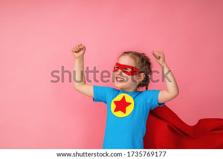 beautiful little girl in a superhero costume, in a red Cape and mask shows how strong she is, isolated against a pink background. Cute kid playing superhero. The concept of Power girl.