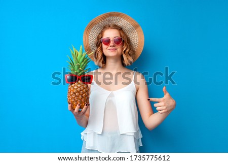 young girl in a sun hat and glasses shows a finger at a pineapple on a blue isolated background, woman tourist in a resort, summer vacation concept