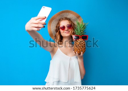 young girl in a sun hat and glasses takes a selfie with pineapple on a blue isolated background, woman tourist taking pictures on vacation