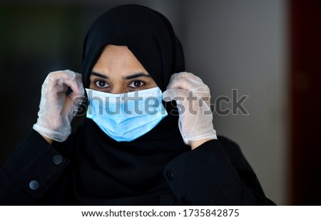 An arab woman wears an face mask respirator while leaning on a shopping cart outside of a Supermarket. The woman is protecting herself from coronavirus and other airborne particles and diseases. Royalty-Free Stock Photo #1735842875