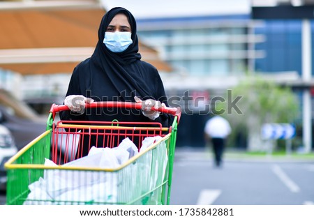 A arab Muslim woman wears an face mask respirator while leaning on a shopping cart outside of a Supermarket. The woman is protecting herself from coronavirus and other airborne particles and diseases. Royalty-Free Stock Photo #1735842881