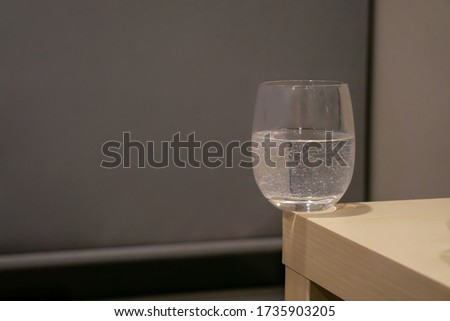 A glass of water with bubbles inside is on the corner of light yellow wooden table in the grey room. A cup on the edge looks dangerously. Object picture with copy space.
