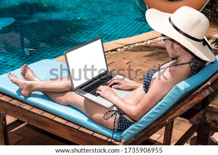 Asian young beautiful woman luxury enjoying relaxing sitting deck chair edge poolside she's working on laptop computer, swimming pool blue water on holiday time