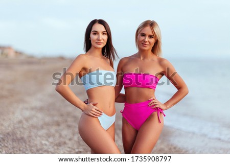 Two amazing tanned girls on the beach, blonde and brunette, long hair, perfect figures.