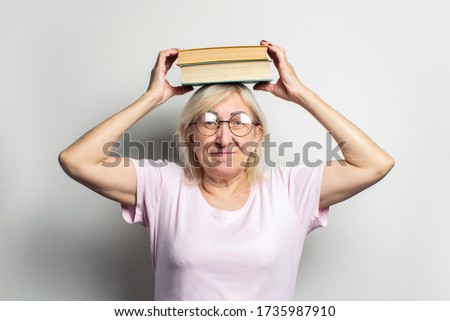 Portrait of an old friendly woman with a smile in a casual t-shirt and glasses holds two books on her head on an isolated light background. Emotional face. Concept book club, leisure