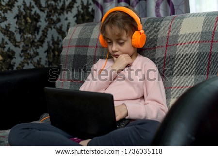 Cute little girl sitting in headphones and with a laptop on the sofa in the room. The concept of distance learning during quarantine.