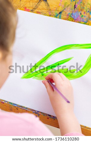 Child's hand drawing green leaf by watercolors on white sheet of paper on an easel. Earth day concept.