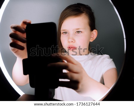 Small girl using camera of smartphone in front of ring light, shooting video for blog. Adorable child learning new technology. 