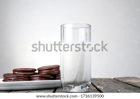 Glass cup of milk in the center on a wooden table and cookies in chocolate icing on a white plate