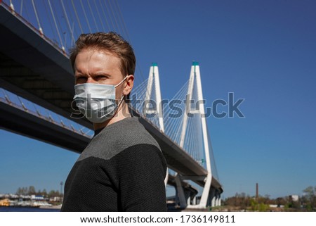 A young man with blond hair stands against the background of a cable-stayed engineering bridge in a medical mask, trying to remove it from her face, social distance
