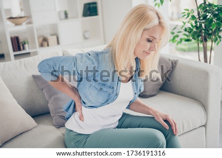 Closeup profile photo of unwell displeased blond mature lady sit couch stay home quarantine hold hand on spine suffer pms touch back massaging injured pain place living room indoors