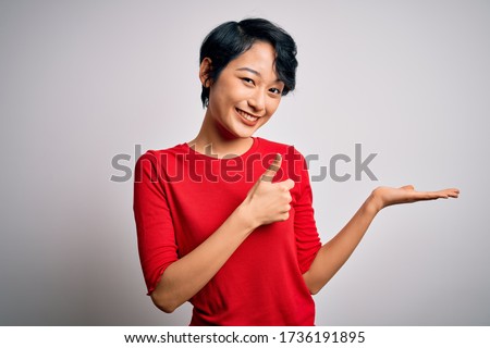 Young beautiful asian girl wearing casual red t-shirt standing over isolated white background Showing palm hand and doing ok gesture with thumbs up, smiling happy and cheerful