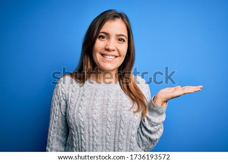 Beautiful young woman wearing casual wool sweater standing over blue isolated background smiling cheerful presenting and pointing with palm of hand looking at the camera.