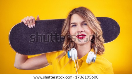 Happy lady in casual yellow polo with red lips and headphones holding skateboard on shoulder and looking at camera while standing on yellow background