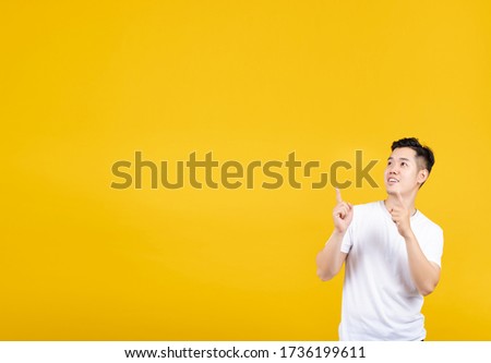 Attractive portrait happy young asian man pointing showing looking side for banner with copy space wearing white t-shirt on yellow background isolated studio shot.