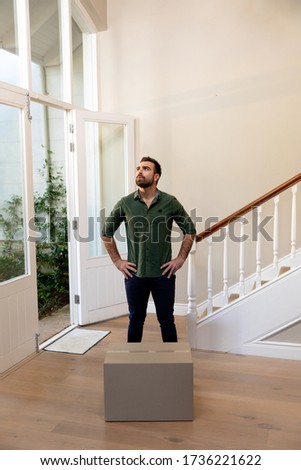 Front view of a Caucasian man arriving at his new home, standing in the hallway looking around with his hands on his hips, a cardboard packing box on the floor in front of him