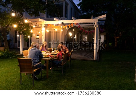 Side view of a multi-generation Caucasian family sitting in the garden outside their house at a dinner table in the evening for a celebration meal together, talking and eating Royalty-Free Stock Photo #1736230580