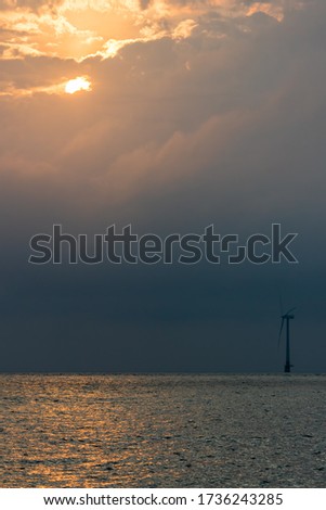 Standing against climate change. Solitary offshore wind turbine landscape representing green energy and global warming. Single windfarm turbine at dawn or dusk with sunlight reflecting off the sea.