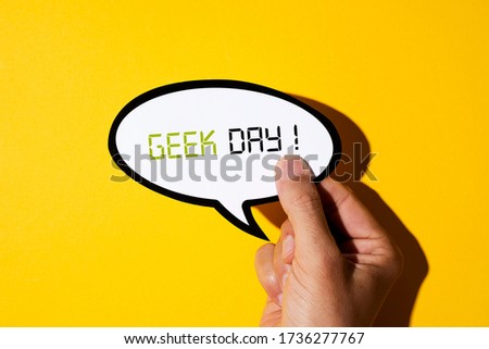 closeup of the hand of a young caucasian man showing a speech balloon, with the text geek day written in it, on a yellow background