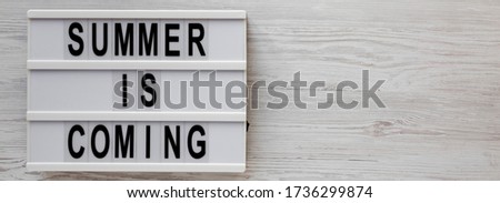 'Summer is coming' text on a lightbox on a white wooden background, top view. Flat lay, overhead, from above. Copy space.