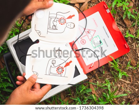 Electrode for attaching to the patient's chest In the hands of people There is a red AED in the back. Placed on the grass. There is a copy space concept, life support equipment, emergency vehicles. Royalty-Free Stock Photo #1736304398