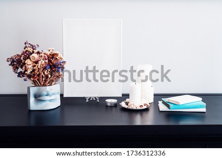 Interior design of room, blank white canvas for artist's mock-up, lips shaped vase with dry flowers bouquet, candles, white books on black table and white wall. Minimalism style.