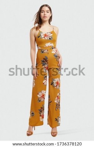 A romantic brown-haired lady in high heel sandals is wearing figure-flattering jumpsuit with thin shoulder straps and wide trouser legs. The mustard jumpsuit is decorated with colorful flowers print. Royalty-Free Stock Photo #1736378120