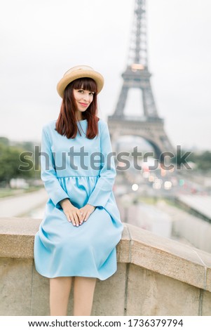 Outdoor image of gorgeous young woman in hat and blue dress sitting on stairs in beautiful European city. Paris, France, Eiffel tower on the background. Travel and people concept