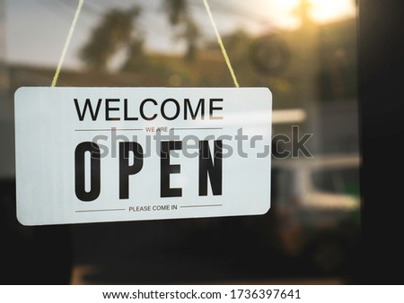 The signage welcomes we are open on a white acrylic sign hanging on the glass door in front of the restaurant. After closing because of Coronavirus and vintage solar situation, symbolic concepts.
