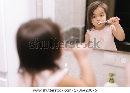 Cute beautiful little girl is brushing her teeth with tooth brush in front of the bathroom mirror, looking at the camera.