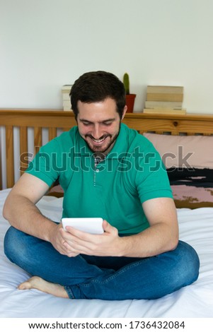 Photo of a young and attractive man with beard reading an ebook and smilling while sitting on his bed.