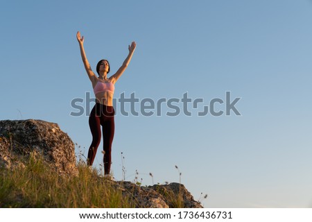Beautiful girl in sportswear remains on top of a mountain and looks at the horizon with a rasied hands, blue sky copyspace on the right side
