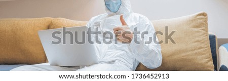 cropped view of man in hazmat suit and protective mask showing thumb up while sitting on sofa and using laptop, panoramic shot