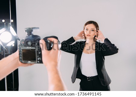 Selective focus of elegant model with hands near hair posing at photographer with digital camera in photo studio