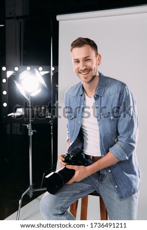 Smiling photographer holding digital camera near floodlight in photo studio