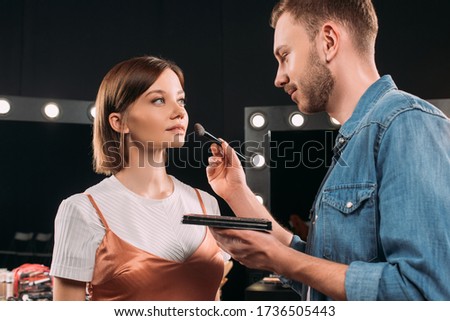 Handsome makeup artist holding makeup set and cosmetic brush near beautiful young woman in photo studio