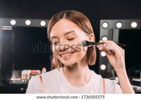 Beautiful smiling woman applying face powder with cosmetic brush in photo studio