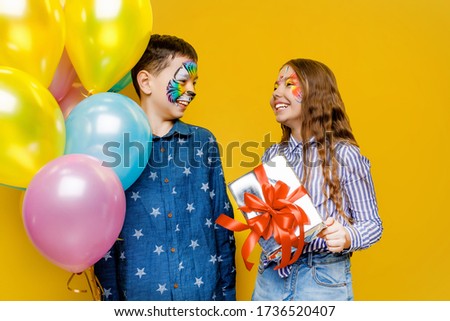 Photo of smiling boy with tiger faceart holding present box and colorful balloons for cute girl isolated on yellow background. Birthday concept.