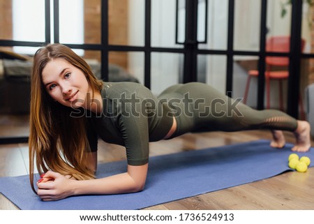 Portrait of young woman working out at home in living room, doing yoga or pilates exercise on blue mat, standing in plank pose (phalankasana).