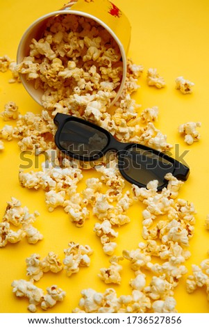 Popcorn spilled out of the bowl. Glasses for viewing the movie are on a yellow background