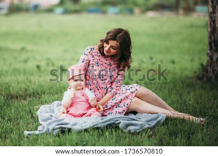 Young beautiful mother with small child have rest in the park on the lawn on warm day in pink clothes under the open sky.