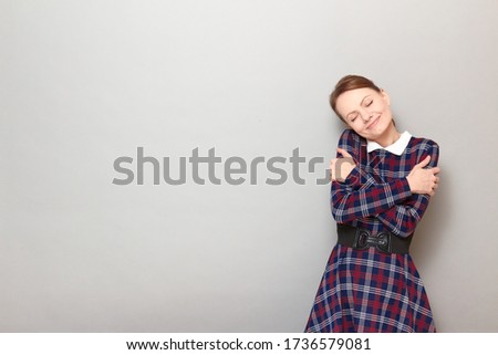 Studio portrait of cute happy girl wearing checkered dress, embracing herself, smiling romantically, with closed eyes, imagining something in mind, standing over gray background, copy space on left