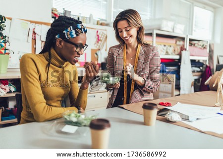 Two young entrepreneur women, or fashion designers working in atelier and making short breakfast break at work